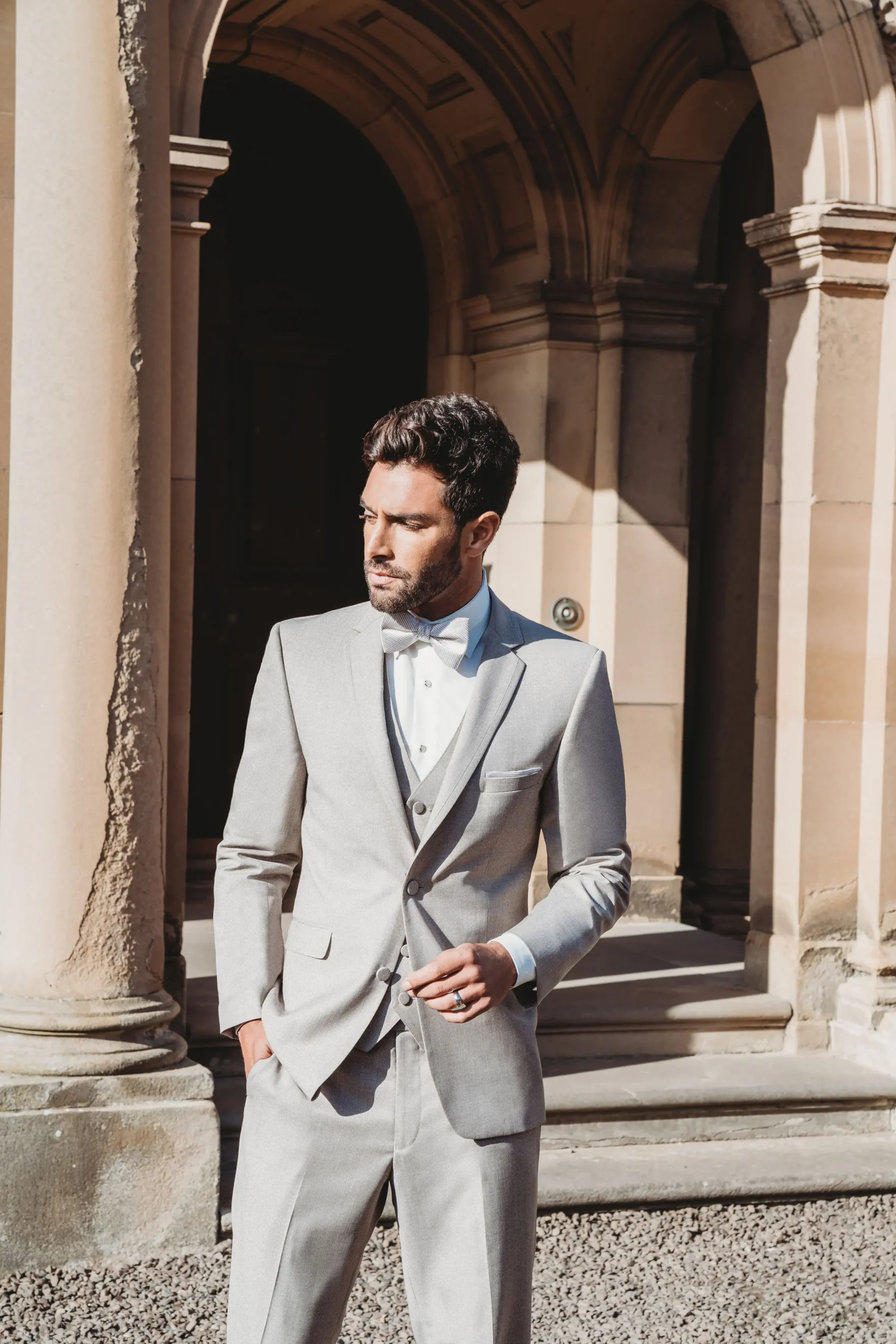 Traje gris perla Allure Men de tres piezas con chaleco, camisa celeste, corbatín beige y solapa notch, medio cuerpo frente a arquitectura clásica — Masculino.do