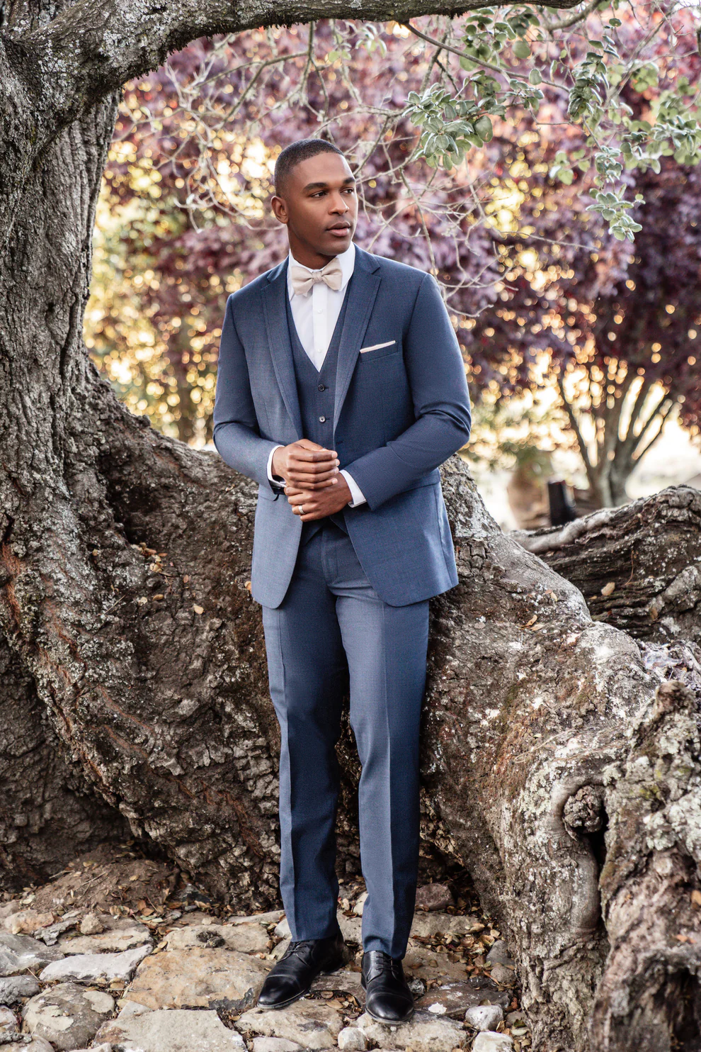Traje azul denim tres piezas con corbatín beige, cuerpo completo apoyado en árbol, ambiente natural — Masculino.do