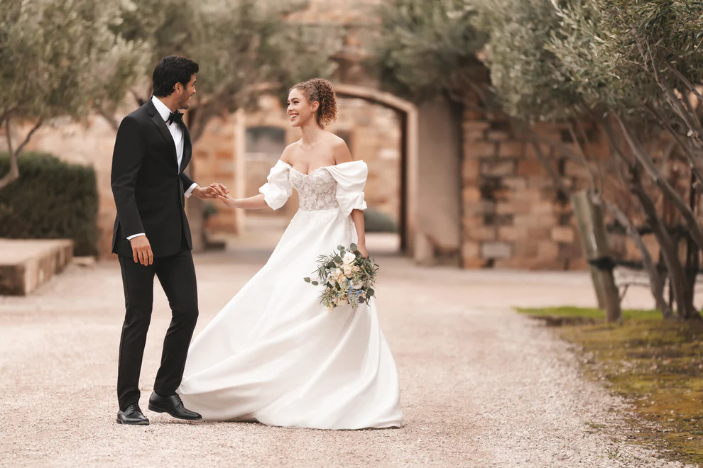 Novio con Tuxedo Milan negro solapa Peak satín y corbatín negro tomado de la mano con novia vestido blanco off-shoulder en jardín de hacienda — Masculino.do