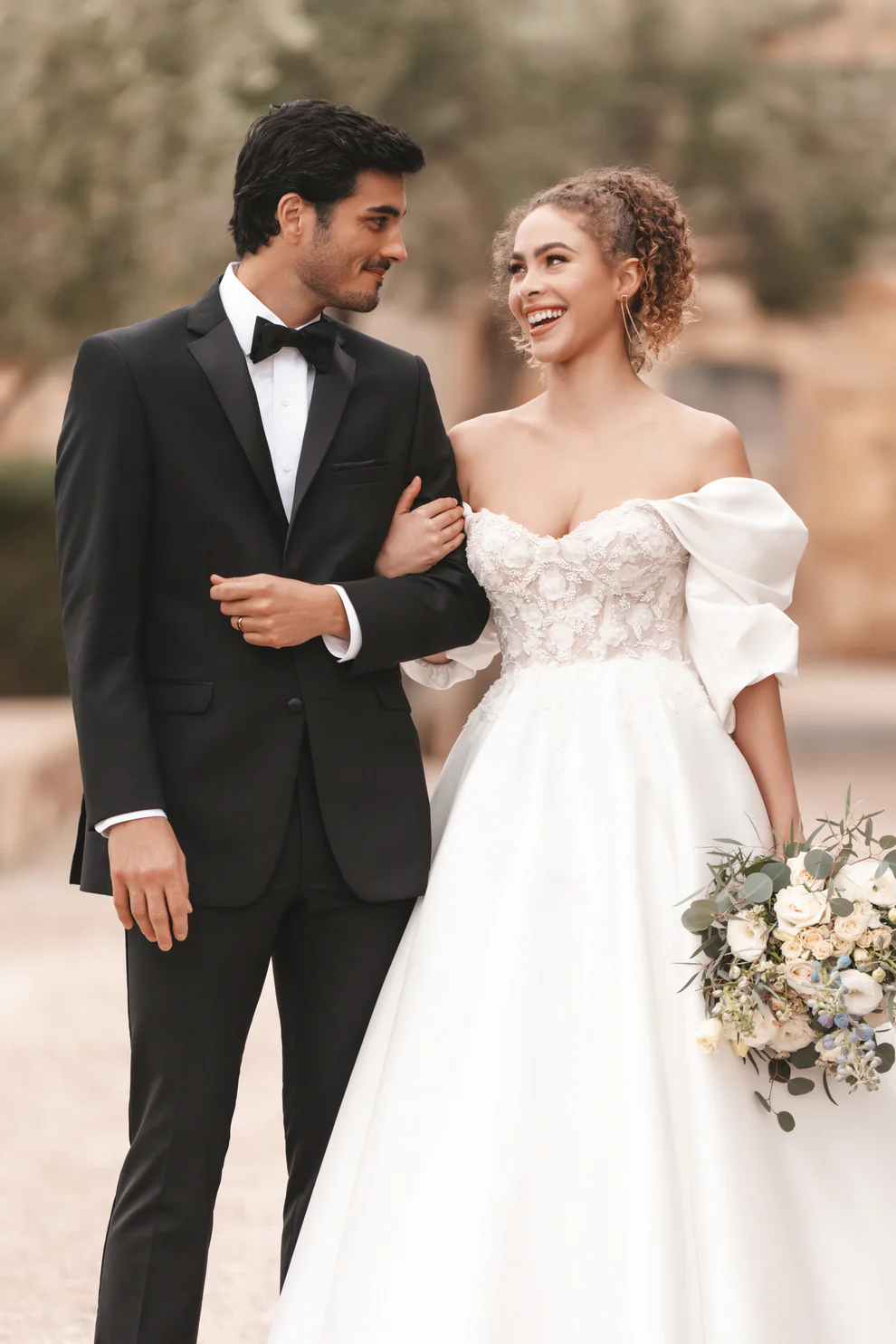 Novio con Tuxedo Milan negro solapa Peak en satín y corbatín negro junto a novia con vestido blanco floral off-shoulder — look de boda al aire libre — Masculino.do