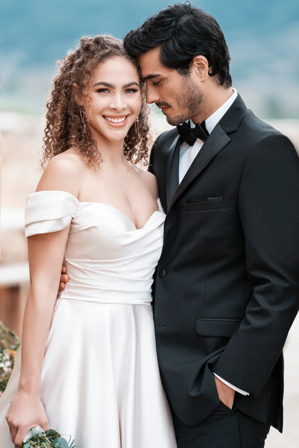 Novio con Tuxedo Milan negro solapa Peak satín y corbatín negro junto a novia con vestido blanco off-shoulder — primer plano romántico boda al aire libre — Masculino.do