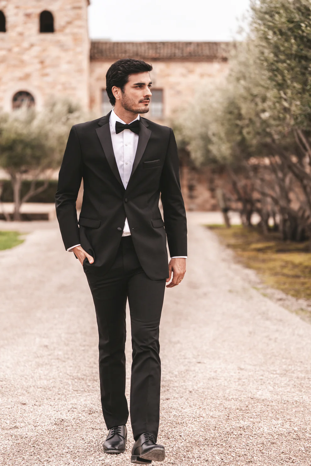 Novio con Tuxedo Milan negro solapa Peak en satín, corbatín negro y camisa blanca caminando por camino de hacienda — look de novio elegante exterior — Masculino.do
