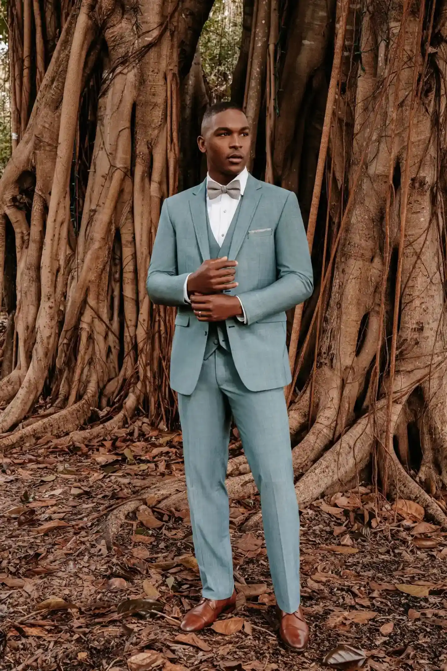 Traje verde sage de tres piezas de cuerpo completo con chaleco, corbatín gris, pañuelo de bolsillo y zapatos café, escenario tropical con raíces — Masculino.do