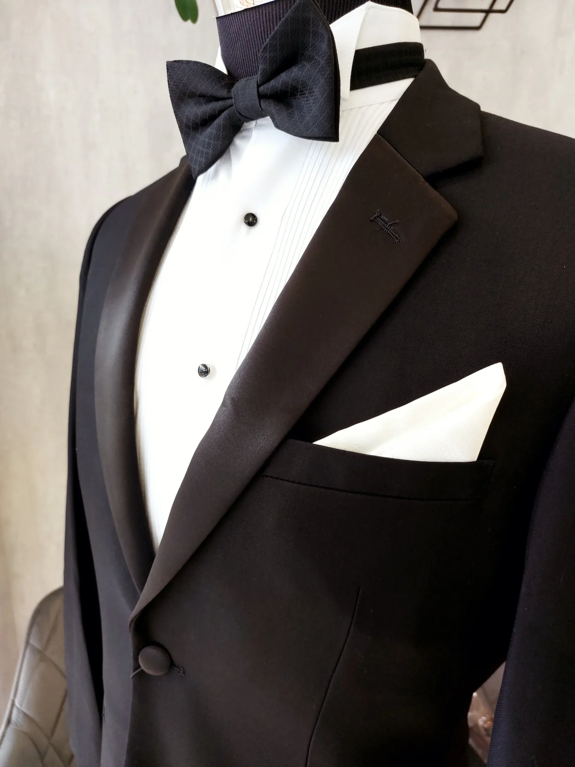 Detalle de esmoquin negro en lana fina con solapa peak en satín, camisa de pechera plisada, corbatín negro y pañuelo blanco en maniquí — Masculino.do