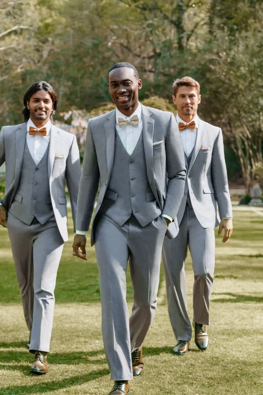 Tres caballeros caminando con trajes grises claros de tres piezas, corbatines en tonos dorado y crema, zapatos café, en jardín — renta de cortejos en Masculino.do